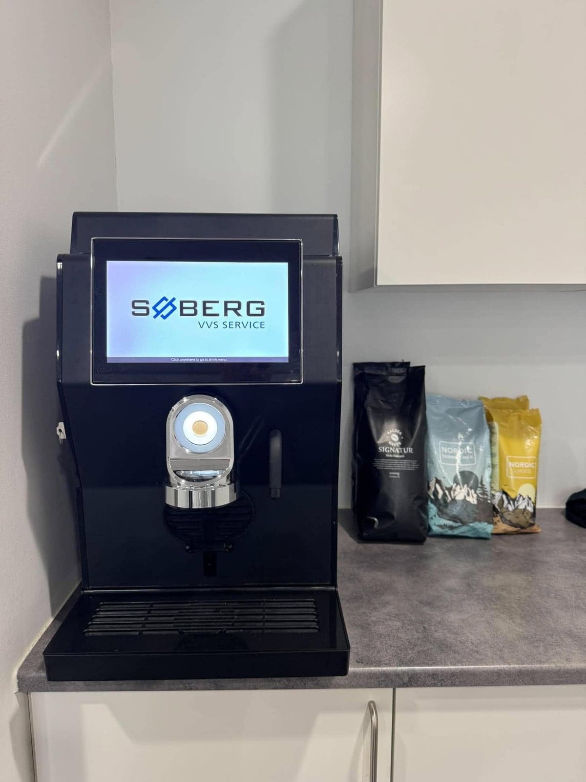 Sort kaffeautomat med touchskærm, der viser logoet for Søberg VVS Service, på et køkkenbord ved siden af poser med Kaldea Signatur, Nordic mælk og Nordic choco