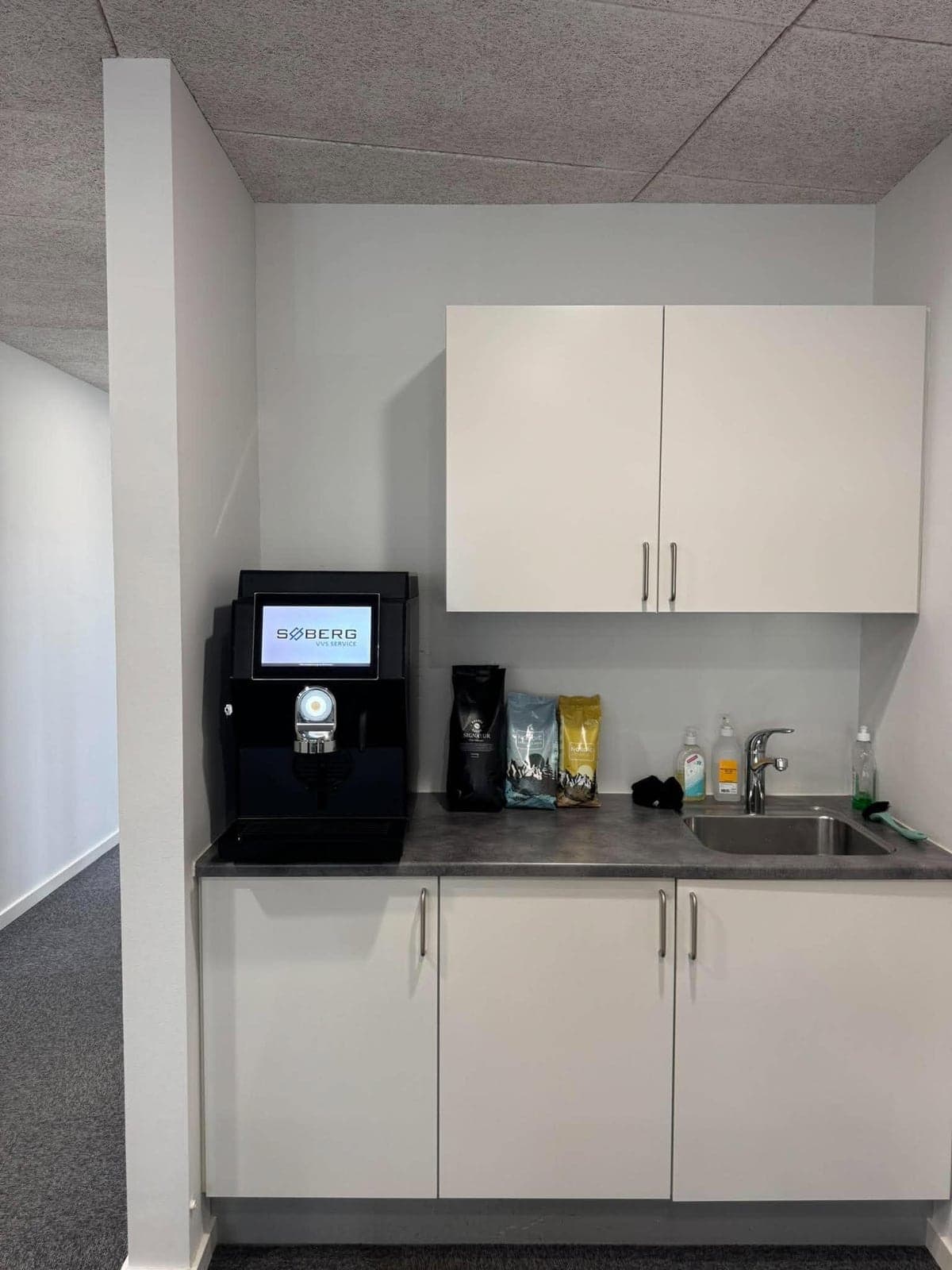 Kontorkøkken med hvide skabe, vask og kaffeautomaten på bordet ved siden af poser med kaffe, mælk og kakao