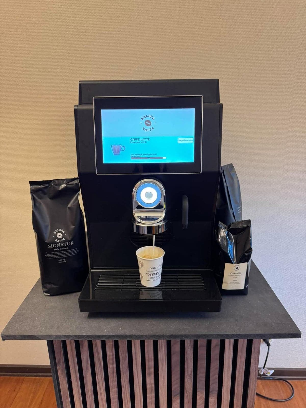 Sort Rhea rhTT1.v+ med touchskærm, der viser Kaldea-logoet, laver en caffe latte i et papkrus. Ved siden af står poser med Kaldea Signatur og Colombia