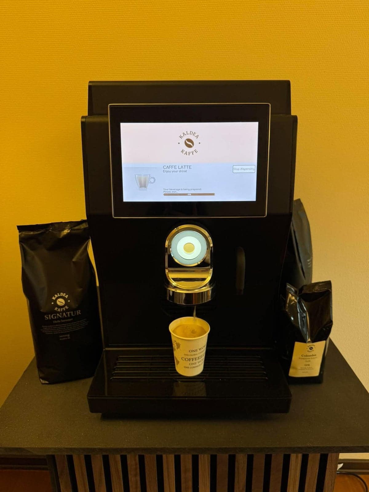 Rhea rhTT1.v+ set forfra, mens den laver en caffe latte, med en pose Kaldea Signatur på den ene side og Colombia på den anden